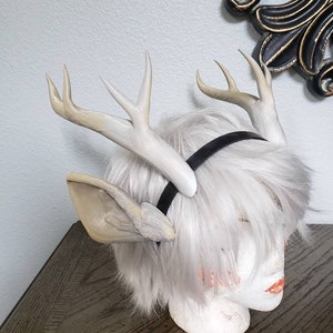 May include: White faux deer antlers headband with a black elastic band. The headband is on a white mannequin head with white hair.