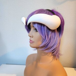 May include: White plastic horns headband with a black elastic band. The headband is worn on a purple wig.