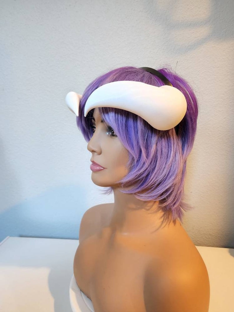 May include: White plastic horns headband with a black elastic band. The headband is worn on a purple wig.