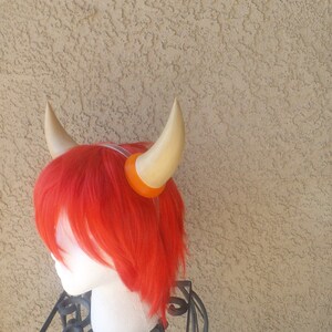 May include: A pair of light brown horns with orange tips, worn as a headband. The horns are made of a soft, flexible material and are designed to be comfortable to wear.