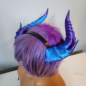 May include: A pair of iridescent blue and purple demon horns attached to a black headband. The horns are made of a soft, flexible material and have a textured surface.