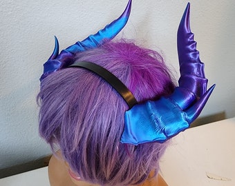 May include: A pair of iridescent blue and purple demon horns attached to a black headband. The horns are made of a soft, flexible material and have a textured surface.