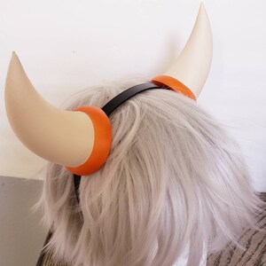 May include: A headband with two white horns with orange tips. The headband is black and the horns are attached to the headband with orange rings.