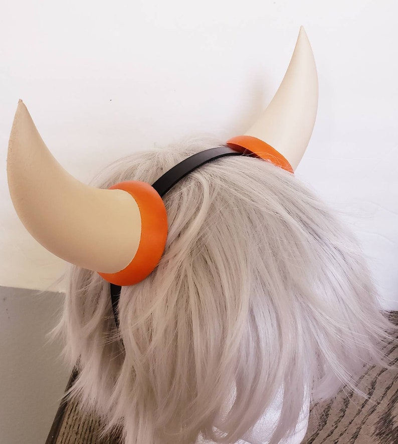 May include: A headband with two white horns with orange tips. The headband is black and the horns are attached to the headband with orange rings.