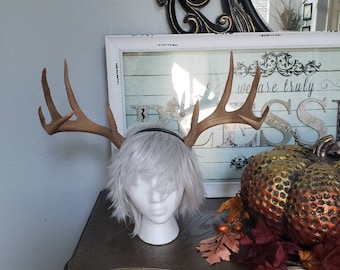 May include: A pair of brown faux deer antlers on a headband, worn by a white wig on a mannequin head. The headband is black. The mannequin is on a dark brown wooden table with a decorative pumpkin and fall leaves.