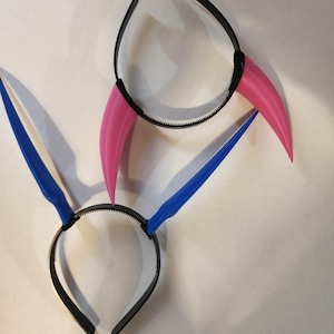 May include: Two black plastic headbands with attached horns. One headband has pink horns and the other has blue horns.