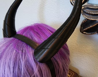 Könnte beinhalten: Ein Paar schwarze, gebogene Hörner aus Kunststoff, die an einem schwarzen Stirnband befestigt sind. Die Hörner sind so konzipiert, dass sie wie Dämonenhörner aussehen.