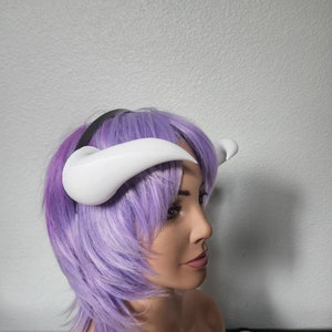 May include: A white headband with two white horns attached to it. The headband is worn by a mannequin with purple hair.