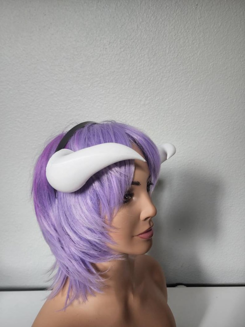 May include: A white headband with two white horns attached to it. The headband is worn by a mannequin with purple hair.