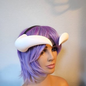 May include: White plastic headband with two curved horns, worn by a mannequin with purple hair.