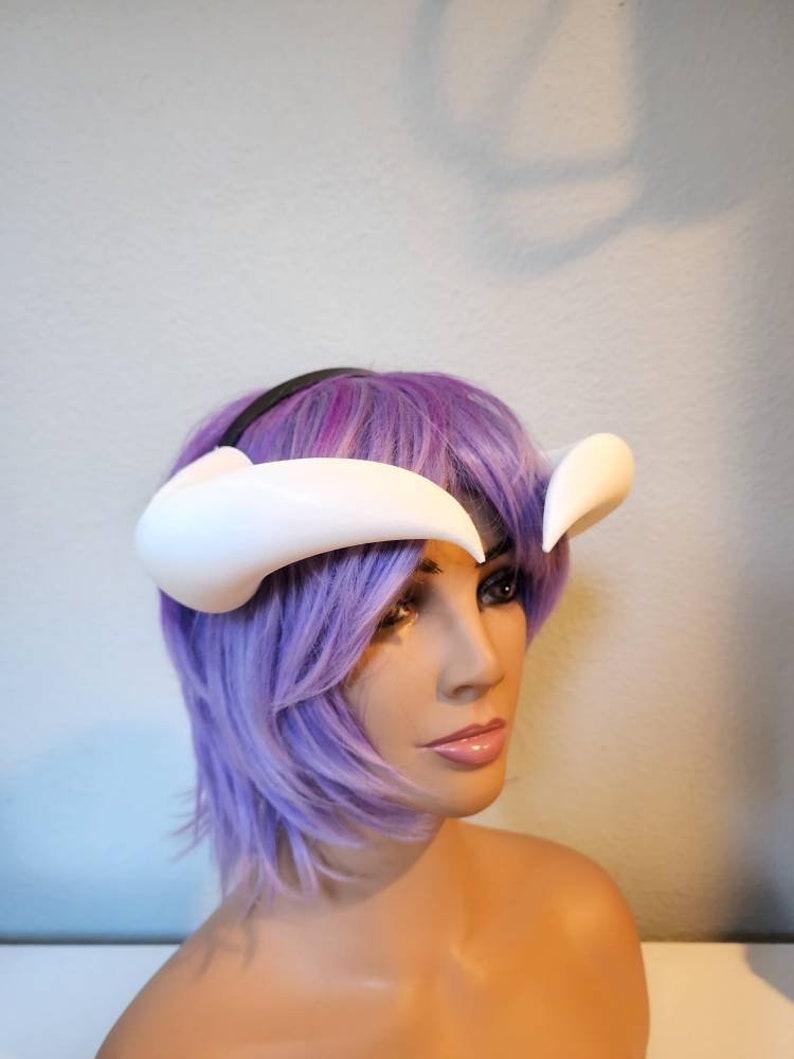 May include: White plastic headband with two curved horns, worn by a mannequin with purple hair.