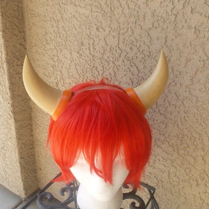 May include: A red wig with two tan horns attached to it. The horns have orange bands around the base. The wig is on a white mannequin head, which is sitting on a black metal stand.