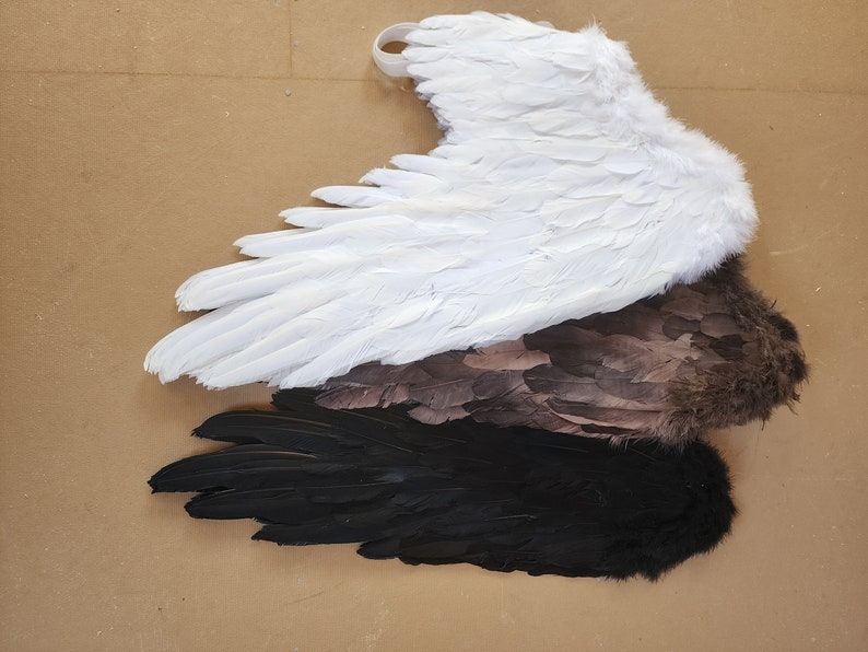 May include: Three pairs of angel wings in different colors: white, brown, and black. The wings are made of feathers and are arranged in a stack.