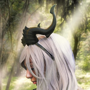 May include: A black headband with a pair of large, curved horns. The horns are detailed with textured scales and have a pointed tip. The headband is worn by a person with long, white hair.