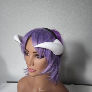May include: A white headband with two white horns attached to it. The headband is worn on a mannequin with purple hair.