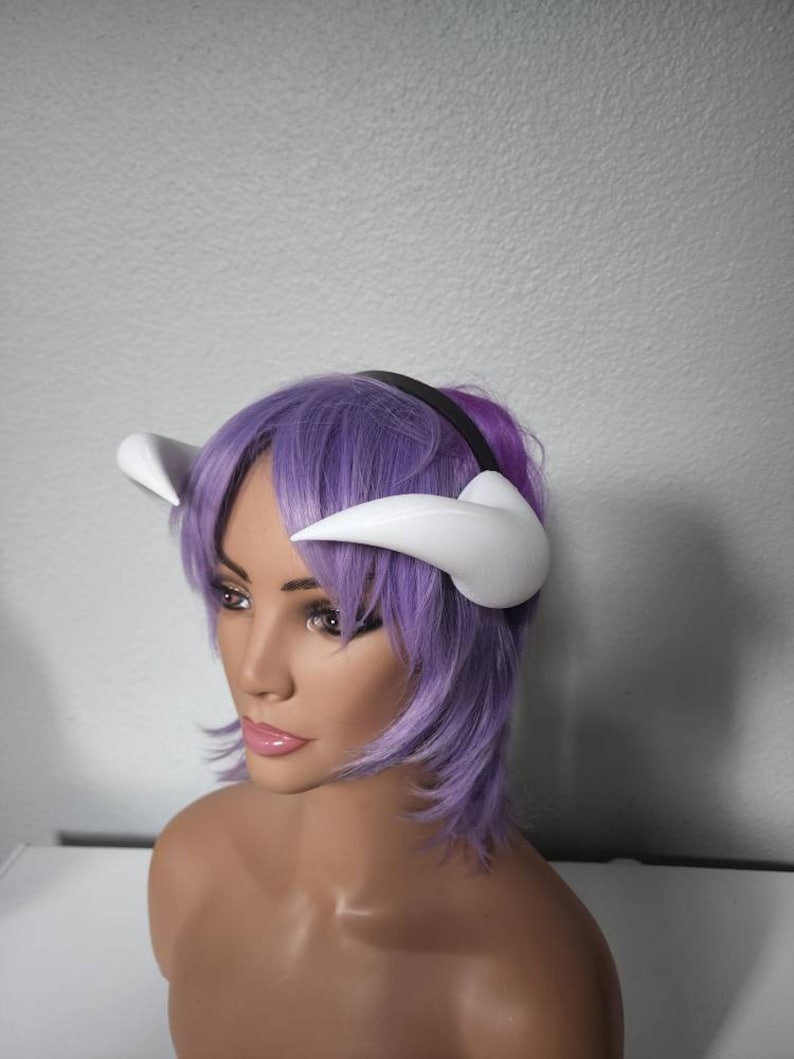 May include: A white headband with two white horns attached to it. The headband is worn on a mannequin with purple hair.