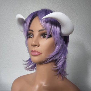 May include: A white plastic headband with two large, curved horns. The headband is worn on a mannequin with a short, purple wig.