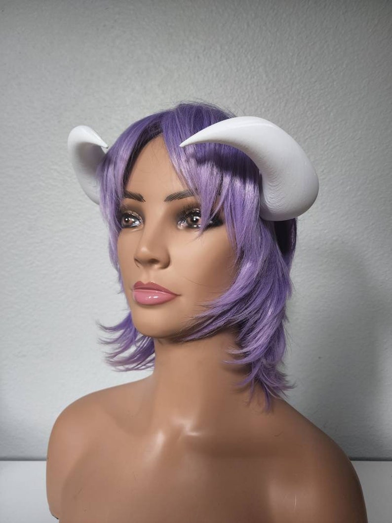 May include: A white plastic headband with two large, curved horns. The headband is worn on a mannequin with a short, purple wig.
