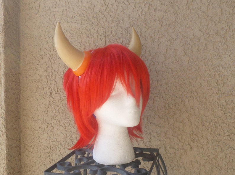 May include: A pair of brown horns with orange bases attached to a red wig on a mannequin head.