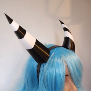 May include: Black and white striped devil horns headband. The headband is made of plastic and is worn on a person with blue hair.