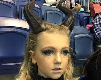 Puede incluir: Una niña joven con cabello rubio que lleva un disfraz negro y un par de cuernos negros. Está sentada en una silla azul con una expresión seria en su rostro.