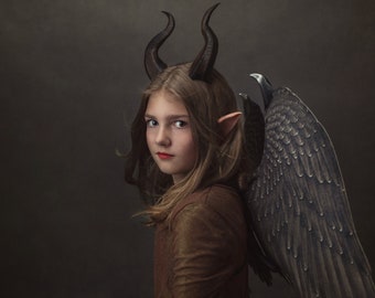 May include: A young person with long brown hair wearing a brown shirt and a black devil costume with horns and wings.