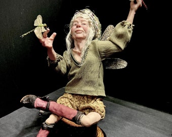 May include: A whimsical fairy doll with long white hair, wearing a green shirt, brown shorts, and pink leggings, sits on a wooden stump. The fairy is holding two dragonfly figurines, one in each hand.