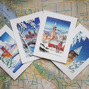 May include: Four winter-themed greeting cards featuring watercolor illustrations of snowy buildings and trees. The cards are white with a white border and are arranged in a square shape.