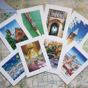 May include: A set of eight greeting cards featuring watercolor illustrations of various scenes. The cards are white with a black border and feature scenes such as a rowing team, a university building, a snowy cityscape, and a library.
