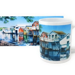 May include: A white ceramic mug with a colorful watercolor illustration of a row of houses on stilts over a body of water. The houses are painted in shades of blue, red, green, and white. The water is a deep blue with reflections of the houses.