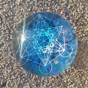 May include: A round, blue glass paperweight with a white geometric design. The design is a complex pattern of lines and circles, resembling a sacred geometry symbol.