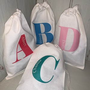 May include: Four white fabric drawstring bags with different coloured letters and names on them. The letters are A, B, C, and D. The names are Albert, Benny, Charlie, and Dolly.