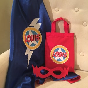 May include: A superhero set comprising a blue cape with a white lightning bolt and a circular emblem with the name "LOUIS", a red tote bag with the same emblem, and a red eye mask.
