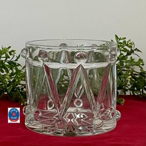 May include: A clear glass jar with a geometric design. The jar has a cylindrical shape with a series of triangular and circular patterns. The jar is sitting on a red surface with greenery in the background.