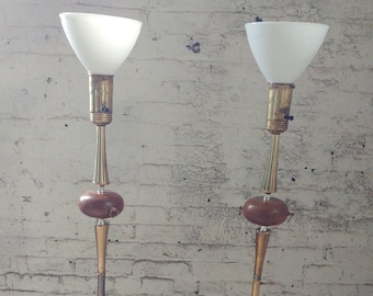 May include: A pair of mid-century modern floor lamps with white shades and wooden bases. The lamps have a brass finish and a unique design with a round, wooden accent piece.