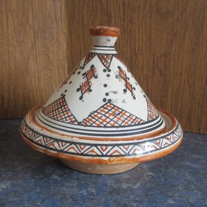 May include: A white ceramic tagine with a conical lid, decorated with geometric patterns in orange and black. The tagine is on a blue tiled surface.