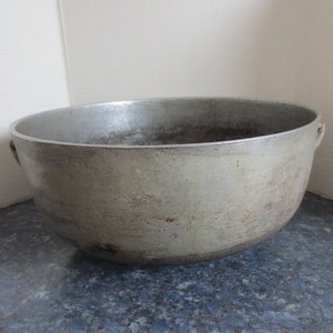 May include: A large, round, silver aluminum bowl with two handles. The bowl has a worn, aged appearance with some rust spots.