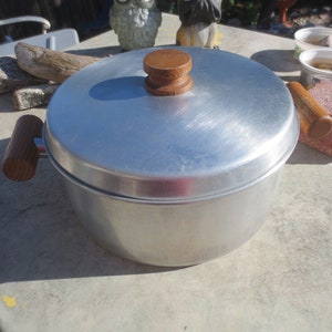 Peut inclure: Une casserole en aluminium argentée avec un couvercle et des poignées en bois. La casserole a un bouton en bois sur le couvercle.