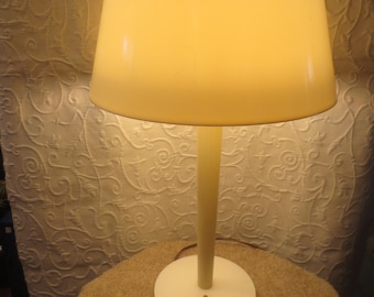 May include: A white table lamp with a white shade and a white base. The lamp is turned on and the shade is illuminated.