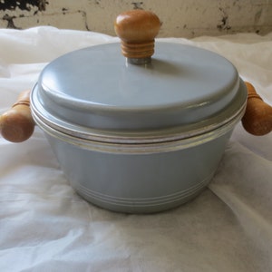 Peut inclure: Une casserole en émail gris avec des poignées en bois et un bouton en bois sur le couvercle. La casserole a une bordure argentée autour du haut.