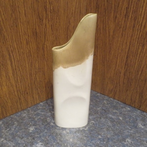 May include: A white ceramic vase with a gold top. The vase has a unique, modern design with a curved shape.