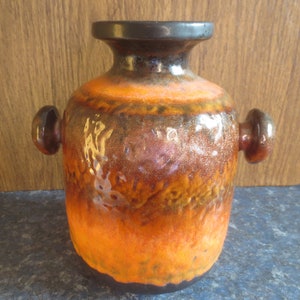 May include: A vintage ceramic vase with a textured surface. The vase features a gradient of colors, transitioning from a dark brown rim to a vibrant orange base. It has two small, rounded handles and a dark brown interior.