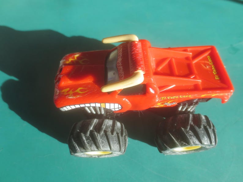 May include: A red toy monster truck with black tires and the text "El Toro Loco". The truck has a bull-like design with horns and a menacing grin. Flames are painted on the sides.
