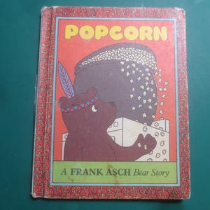 May include: Vintage children's book cover with the title "Popcorn" in yellow letters on a red background. A brown bear with a feather headdress is next to a pot of popcorn. The text "A Frank Asch Bear Story" is below.