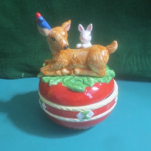 Peut inclure: Boîte à bijoux en céramique avec un cerf et un oiseau bleu et rouge sur l'oreille, et un lapin blanc sur le dos. Le cerf et le lapin reposent sur des feuilles vertes, sur une boîte rouge ronde avec des accents blancs et verts.