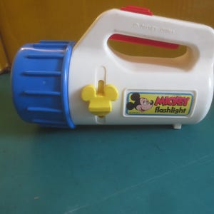 May include: A white plastic Mickey Mouse flashlight with a blue lens and a red switch. The handle has a red accent. The flashlight has a yellow on/off switch and a Mickey Mouse graphic with the words "Mickey Flashlight".