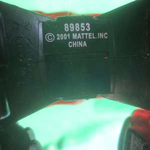 May include: Close-up of a black plastic toy component with the text "89853 &copy; 2001 MATTEL.INC CHINA" in white. The component has a rectangular shape with a slightly curved design. The background is blurred with green and red hues.