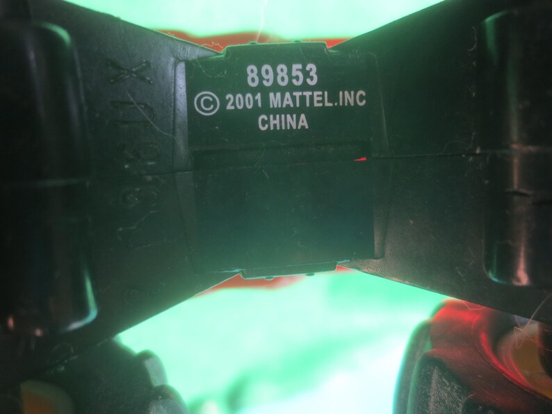 May include: Close-up of a black plastic toy component with the text "89853 &copy; 2001 MATTEL.INC CHINA" in white. The component has a rectangular shape with a slightly curved design. The background is blurred with green and red hues.