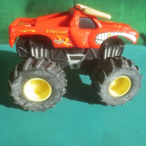 May include: A red toy monster truck with black tires and yellow wheel centers. The truck has a bull horn on top and a cartoonish face with a wide grin. The text "El Toro Loco" is visible on the side.