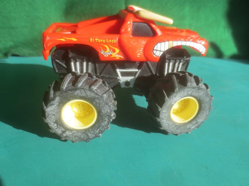 May include: A red toy monster truck with black tires and yellow wheel centers. The truck has a bull horn on top and a cartoonish face with a wide grin. The text "El Toro Loco" is visible on the side.